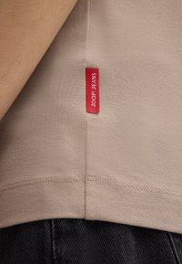 Tissu de T-shirt en coton beige avec une étiquette rouge "JOOP! JEANS" visible cousue au niveau de la couture, associé à un matériau en denim foncé en dessous.