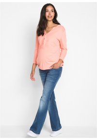 Top maternità color corallo con scollatura henley e tre bottoni, abbinato a jeans a zampa d'elefante blu e sneakers bianche.