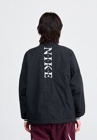 Svart lett jakke med en avslappet passform, som har en dristig hvit "NIKE"-logo på ryggen, teksturert stoff og ståkrage.