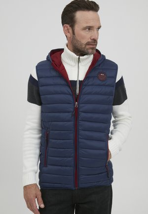 Navy padded vest with a red hood, featuring horizontal quilting, side pockets, and a logo patch. Worn over a white high-neck sweater.