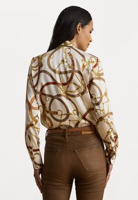 Rakstainā zīda blūze ar zelta ķēdi un sarkaniem akcentiem; valkāta ar brūnajām pieguļošajām biksēm, izceļot dizainu un tekstūru.