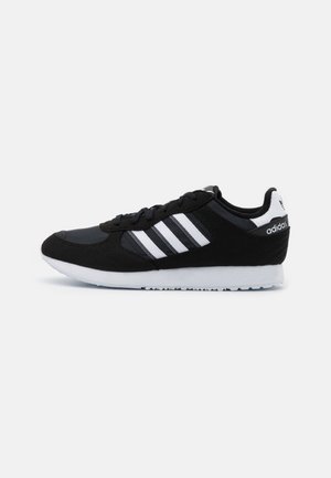 Chaussure de sport noire avec une tige en daim et synthétique. Équipée de trois rayures blanches, d'un laçage à l'avant et d'une semelle en caoutchouc blanche.