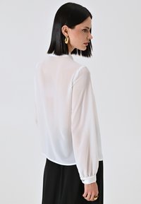 Camicia bianca e trasparente con maniche lunghe e gonfiate e polsini. Texture liscia; orlo curvo; presenta un colletto e una vestibilità comoda. Pantaloni neri visibili sotto.