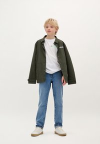 Giacca leggera verde sopra una maglietta bianca, abbinata a jeans azzurri e sneakers bianche; design semplice, vestibilità rilassata, senza loghi visibili.