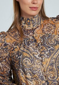 Blusa abotonada de tejido transparente con un patrón de paisley en colores azul marino, dorado y crema; cuello alto y mangas abullonadas.