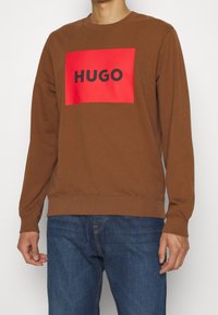 Sweat-shirt marron à col rond, avec un grand patch rectangulaire rouge affichant "HUGO" en lettres noires épaisses. Texture douce, coupe décontractée.