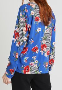Donna che indossa una blusa azzurra a maniche lunghe con fiori rossi, rosa e bianchi e toppe a righe nere e bianche sulla schiena e sulle maniche.