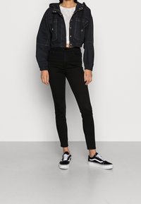 Chaqueta negra corta con capucha, dos bolsillos frontales y puños elásticos, combinada con jeans ajustados negros y zapatillas negras con acentos blancos.