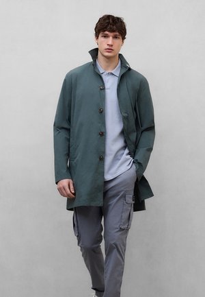 Joven vestido con un abrigo verde abotonado sobre una camiseta tipo polo azul claro y pantalones cargo grises, de pie contra un fondo gris liso.