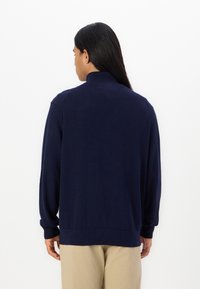 Granatowy sweter z dzianiny z wysokim kołnierzem, długimi rękawami i ściągaczami na nadgarstkach. Miękka textura i luźny krój widoczne z tyłu.