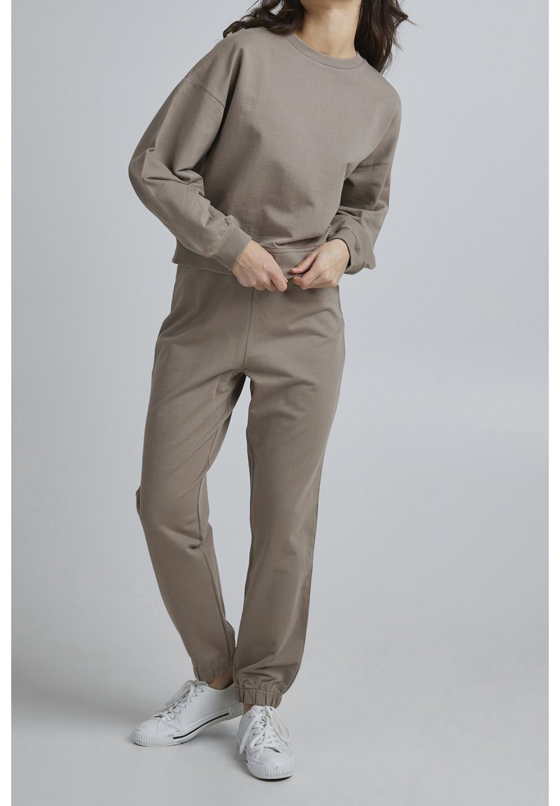 Femme portant un sweat-shirt taupe et un pantalon de jogging assorti avec des baskets blanches, ajustant la taille du pantalon sur fond gris uni.