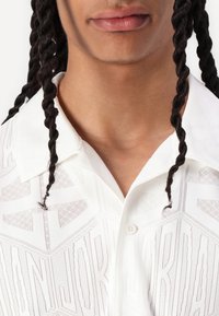 Chemise blanche à manches courtes avec des motifs géométriques et une texture subtile. Elle dispose d'un col pointu et d'une fermeture à boutons.