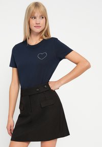 Camiseta de algodón navy con un diseño de corazón hecho de pequeños remaches en el pecho, combinada con una falda negra que presenta dos botones y bolsillos frontales.