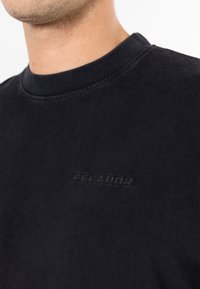 Close-up of a person wearing a black crew neck shirt with subtle embossed text "PEGADOR" on the chest.