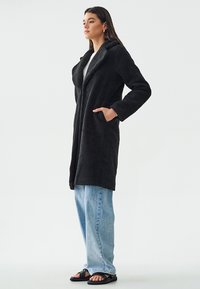 Abrigo de forro negro con cuello, de mangas largas y bolsillos laterales. Llevado sobre una camiseta gris y combinado con jeans azul claro y sandalias negras.