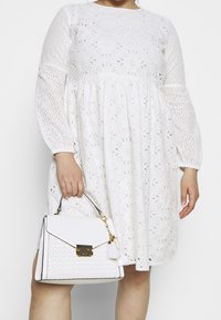 Bíl dress s vyšívaným očkovým vzorem, dlouhými rukávy a nařasenou sukní; v kombinaci s bílou strukturovanou taškou s třásní.