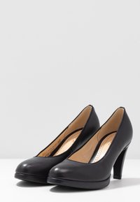 Pár klasických černých kožených dámských bot na vysokém podpatku s kulatou špičkou na bílé ploše a šedém pozadí.