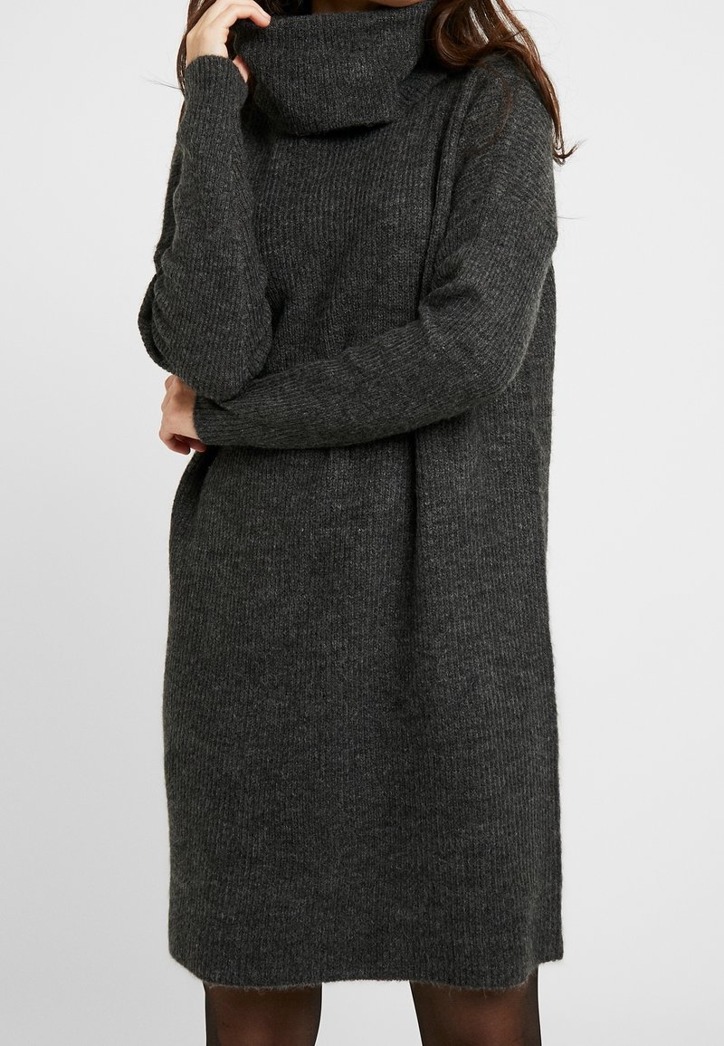 Femme portant une robe-pull en tricot côtelé gris foncé à manches longues avec un grand col roulé.