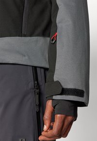 Chaqueta resistente al agua con una combinación de colores negro y gris. Las características incluyen un bolsillo con cremallera, puños de Velcro y un acabado de tela texturizada.