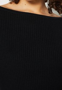 Nahaufnahme einer Person, die ein schwarzes, geripptes, schulterfreies Oberteil trägt, das den Hals, das Schlüsselbein und lockige Haarsträhnen zeigt.