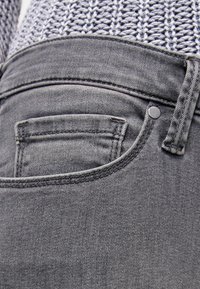 Harmaat farkut, joissa on viiden taskun muotoilu, pieni takatasku ja metallikoriste. Pinta on sileä, ja siinä on hienovarainen haalistuma.