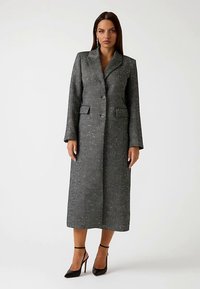 Long manteau gris en tissu texturé, avec deux poches avant, des revers crantés et une fermeture à un seul bouton, porté avec des escarpins noirs.