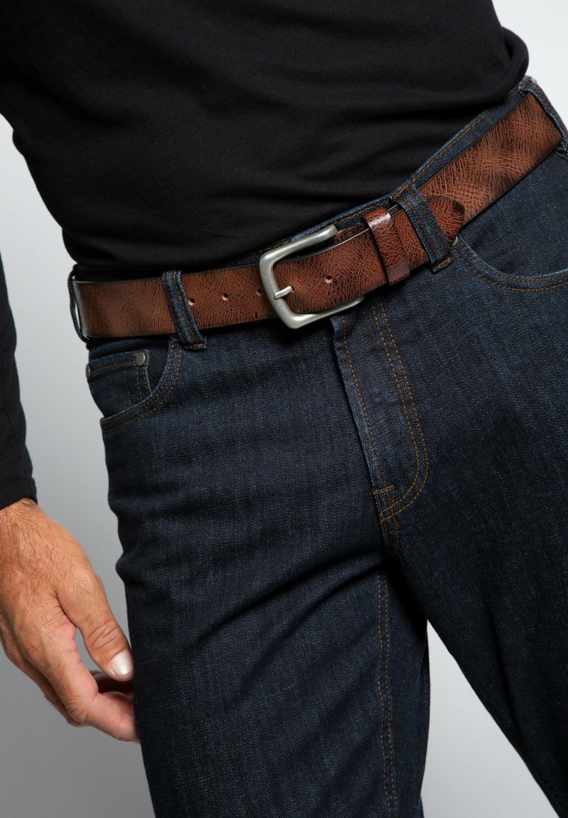 Ceinturon en cuir marron avec une boucle rectangulaire en argent, porté sur un jean en denim bleu foncé. Le jean présente des coutures contrastées et une poche latérale.