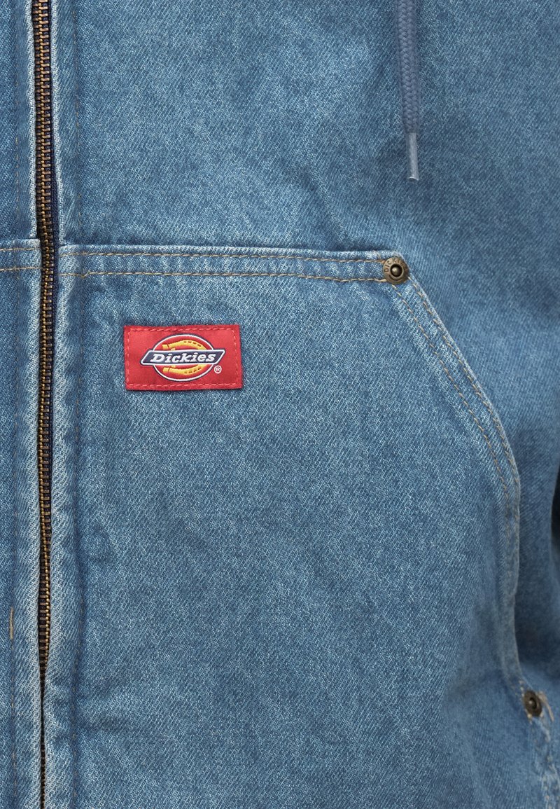 Jeansjacke in Hellblau mit braunem Reißverschluss und rotem Dickies-Logo-Patch auf der Brust. Verfügt über zwei Fronttaschen und sichtbare Nähte.
