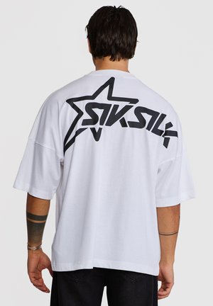 Mann trägt weißes, übergroßes T-Shirt mit schwarzem Stern und stilisiertem "SIKSILK"-Logo auf dem oberen Rücken, vom Betrachter abgewandt.
