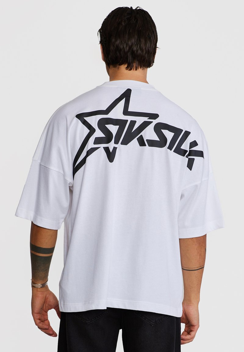Άνδρας που φοράει λευκό, oversized T-shirt με μαύρο αστέρι και στυλιζαρισμένο λογότυπο «SIKSILK» στην πάνω πλάτη, κοιτάζοντας μακριά από την κάμερα.