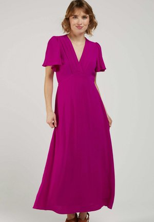 Femme aux cheveux courts portant une robe maxi magenta à encolure en V avec des manches courtes flottantes, debout devant un fond clair uni.