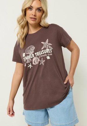 Blondynka ubrana w brązowy T-shirt "Summer Treasures" z nadrukami muszli i rozgwiazd, zestawiony z jasnoniebieskimi denimowymi szortami.