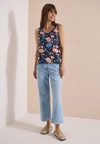Woman smiling wearing a sleeveless navy floral top, light blue wide-leg jeans, and beige slip-on shoes against a beige and white background.