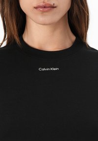 Čierne bavlnené tričko s okrúhlym výstrihom, krátkymi rukávmi a bielym logom Calvin Klein umiestneným na hrudi. Hladká textúra, voľný strih.