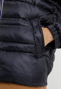 Marinblå puffjacka med ett texturerat, kviltat mönster, som har en sidoficka och elastiska ärmslut för en snug passform.