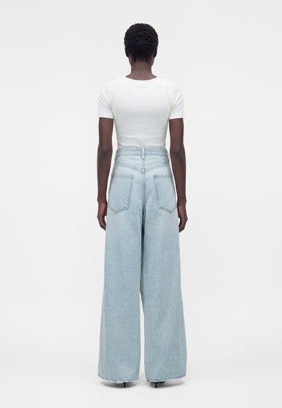 Személy világoskék, magas derekú, széles szárú farmernadrágot és testhez simuló fehér rövid ujjú felsőt visel, háttal áll egy egyszínű háttér előtt.
