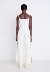 Pantalons larges blancs avec des rayures noires sur les côtés, fabriqués à partir d'un tissu doux. Associés à un débardeur blanc ajusté. Vue de derrière.