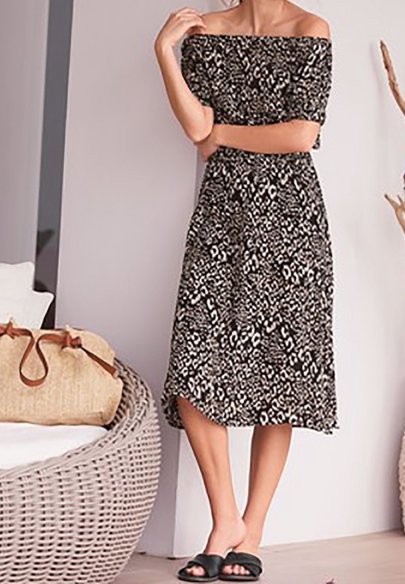 Femme portant une robe midi à imprimé animal noir et blanc, épaules dénudées, debout à l’intérieur près d’une chaise en osier avec un sac beige.