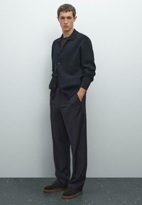Dark navy cardigan with a collared neckline, button-up front, and side pockets, paired with loose-fitting dark trousers and brown shoes.