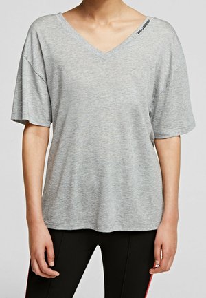 Personne portant un t-shirt ample gris clair à col en V et manches courtes, ainsi qu'un pantalon noir avec une bande rouge sur le côté.
