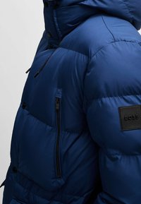 Blue puffer jacket with a hood, featuring a zippered pocket and a leather logo patch. Smooth, shiny finish, and quilted design.