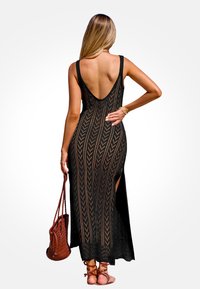 Femme avec de longs cheveux portant une robe maxi en crochet sans manches noire, dotée d'un dos décolleté et d'une fente sur le côté, tenant un sac à main tissé marron.