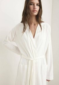 Rochie albă cu mâneci lungi, de tip V, cu talie legată și două buzunare frontale. Material neted cu textură minimă, design simplu.