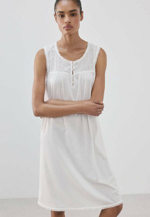 Mujer con los brazos cruzados, vestida con un vestido blanco sin mangas con la parte superior de encaje con ojales y detalles de botones, de pie sobre un fondo claro liso.