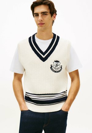 Joven con chaleco de punto blanco con rayas azul marino y logo de Tommy Hilfiger sobre camiseta blanca, con las manos en los bolsillos de unos vaqueros oscuros, frente a un fondo blanco.