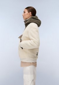 Κρέμα χρώματος fleece μπουφάν με πράσινη κουκούλα, που διαθέτει μπροστινή τσέπη και μαύρες μεταλλικές λεπτομέρειες. Ο ύφασμα έχει μαλακή και υφασμάτινη υφή.