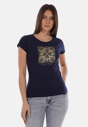Donna che indossa una t-shirt aderente blu navy con motivo geometrico a forma di stella dorata e testo "YES ZEE", abbinata a jeans grigi chiari.