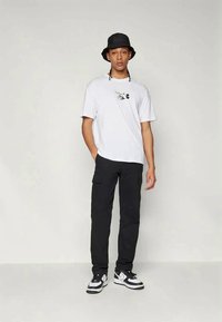 T-shirt branca de manga curta com um estampado gráfico, combinada com calças cargo pretas e sapatilhas em preto e branco; usada com um chapéu de balde preto.