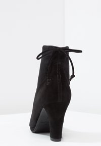 Černá kotníková bota z broušené kůže s vysokým podpatkem, zaoblenou špičkou a zavazováním vzadu. Hladká textura a čisté detaily švů.