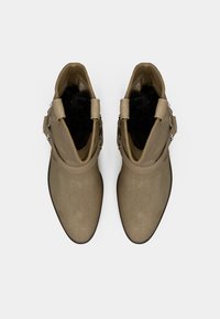 Ζευγάρι μπεζ μποτάκια αστραγάλου από σουέντ με λουριά και ασημένιες τρουκς, σε θέα από πάνω σε λευκό φόντο.